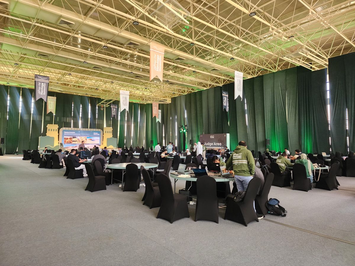 The build hall: round tables of jammers under a high steel roof, a presentation screen at the far end.